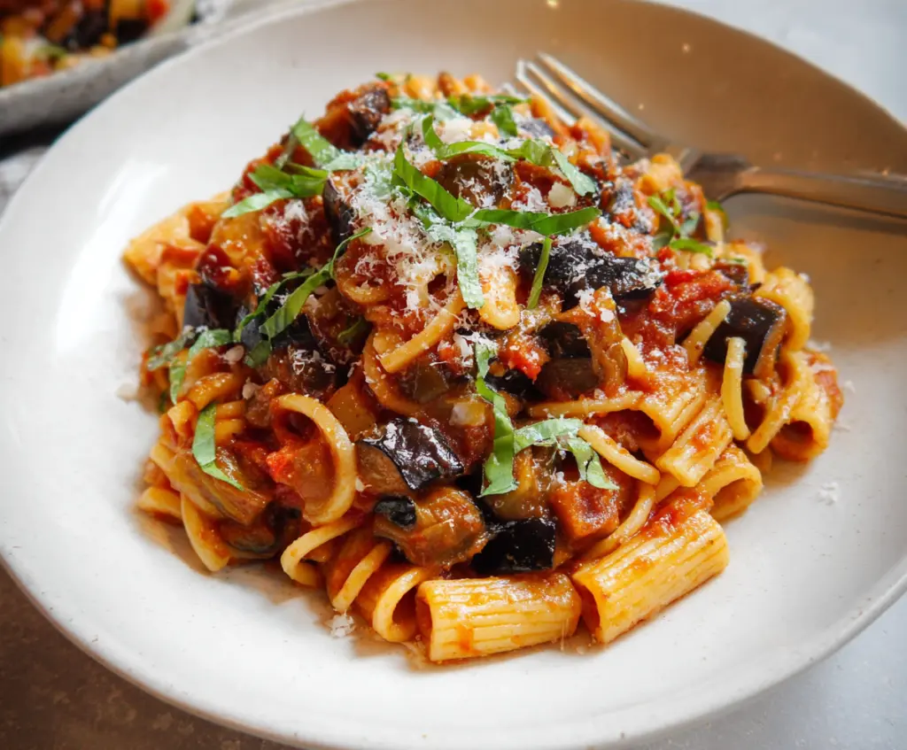 Delicious eggplant pasta dish garnished with fresh herbs on a white plate.